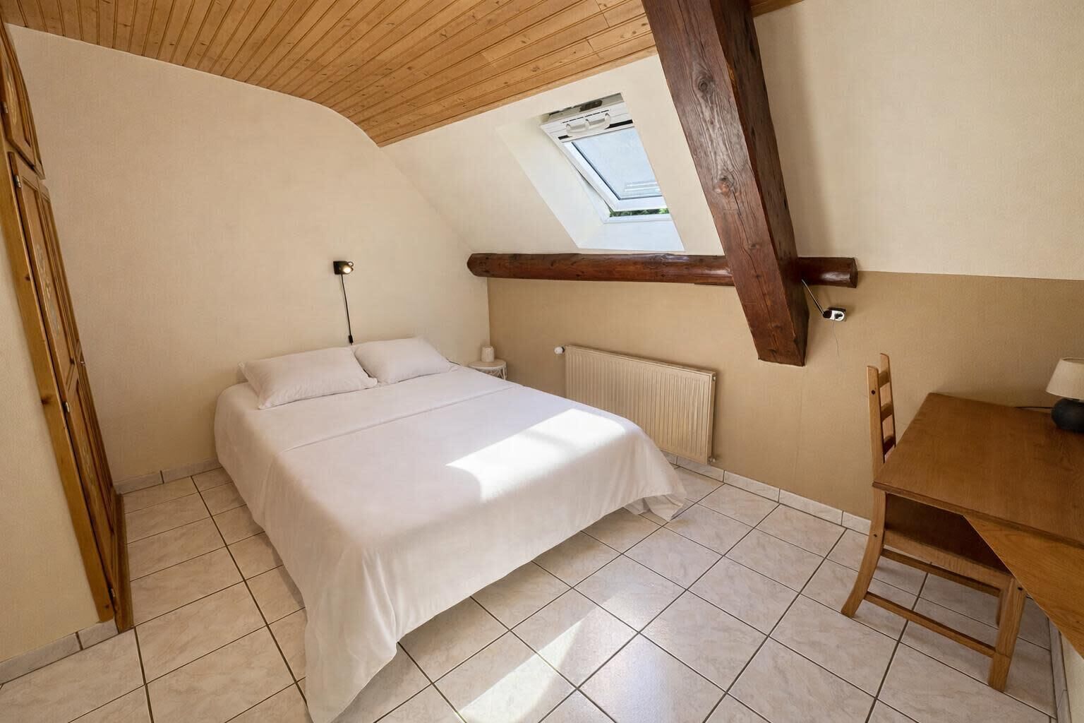Un dormitorio con cama, mesita de noche, mesa, silla, radiador y tragaluz.