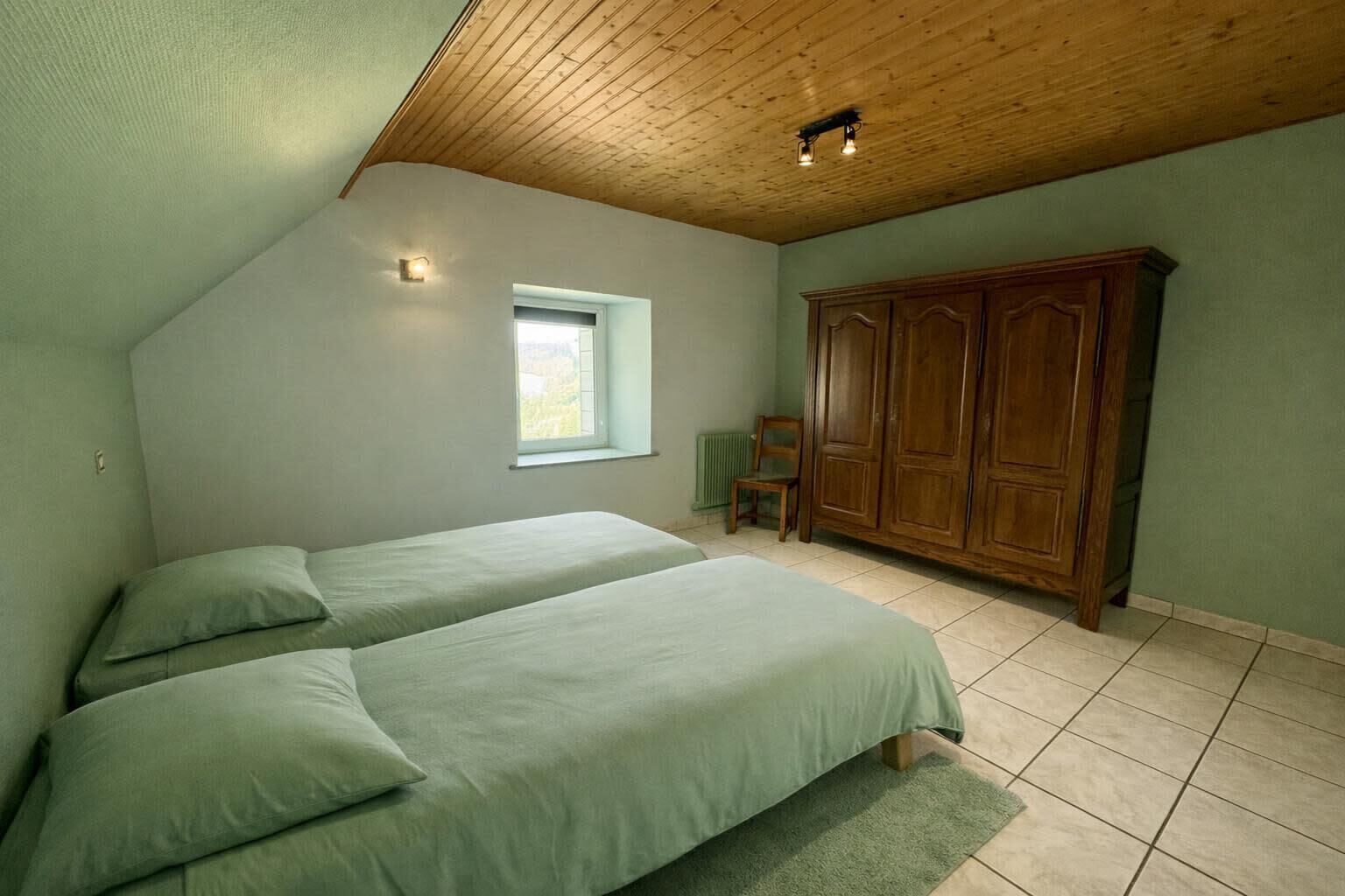 A bedroom with two beds, a window, a wooden wardrobe, and a chair.