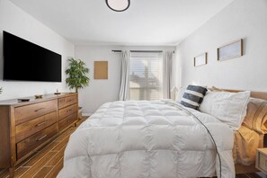 Une chambre à coucher avec un lit, une télévision, une commode, une plante et une fenêtre avec des rideaux.