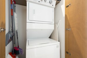 A compact laundry room with a stacked washer and dryer, a mop, and a broom. - Charming Coastal Resort With Ocean Views, Pool, Gym and Shared Amenities (Cabo San Lucas)