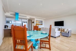 A modern living room with a round glass table, wooden chairs, a sofa, and a TV mounted on the wall. - Charming Coastal Resort With Ocean Views, Pool, Gym and Shared Amenities (Cabo San Lucas)