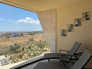 A balcony with a view of a desert landscape, a seating area with chairs, and decorative wall art. - Charming Coastal Resort With Ocean Views, Pool, Gym and Shared Amenities (Cabo San Lucas)