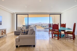 A spacious living area with a sofa, a dining table with chairs, and a view of a cityscape through large windows. - Charming Coastal Resort With Ocean Views, Pool, Gym and Shared Amenities (Cabo San Lucas)