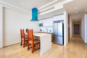 A modern kitchen with white cabinetry, a stainless steel refrigerator, and a wooden dining table with two chairs. - Charming Coastal Resort With Ocean Views, Pool, Gym and Shared Amenities (Cabo San Lucas)