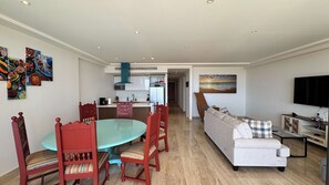 A modern living room with a sofa, a dining table with chairs, a kitchen area, and a TV mounted on the wall. - Charming Coastal Resort With Ocean Views, Pool, Gym and Shared Amenities (Cabo San Lucas)