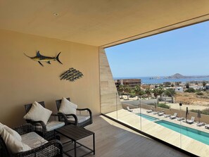 A balcony with wicker furniture, a pool, and a view of the sea and buildings. - Charming Coastal Resort With Ocean Views, Pool, Gym and Shared Amenities (Cabo San Lucas)