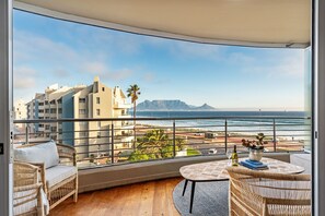A balcony with a view of the ocean and buildings, wicker furniture, and a table with a bottle and book. - Portico 306 (Cape Town)