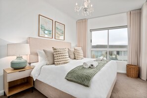 A bedroom with a bed, a nightstand, a lamp, a chandelier, a window with curtains, and artwork on the wall. - Portico 306 (Cape Town)