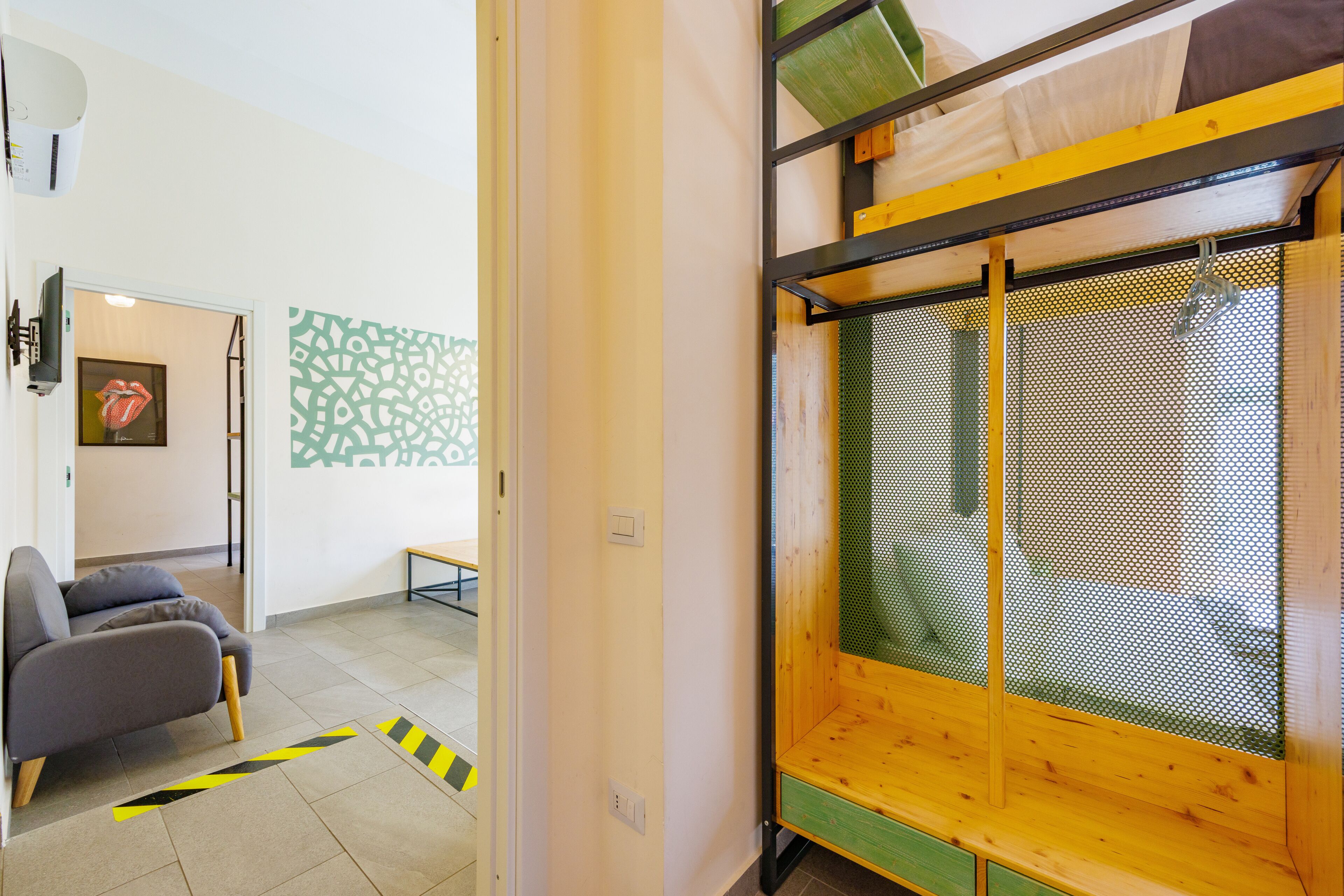 Een kamer met een stapelbed, een grijze bank, een behang met een patroon, en een deur naar een andere kamer.