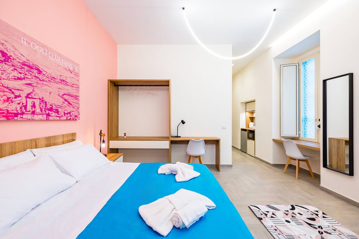 A hotel room with a bed, a desk, a chair, a mirror, and a wall with a pink artwork.