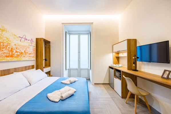 A hotel room with a bed, a desk, a chair, a TV, and a window. - Glow Tripla - Solaris Suites (Naples)