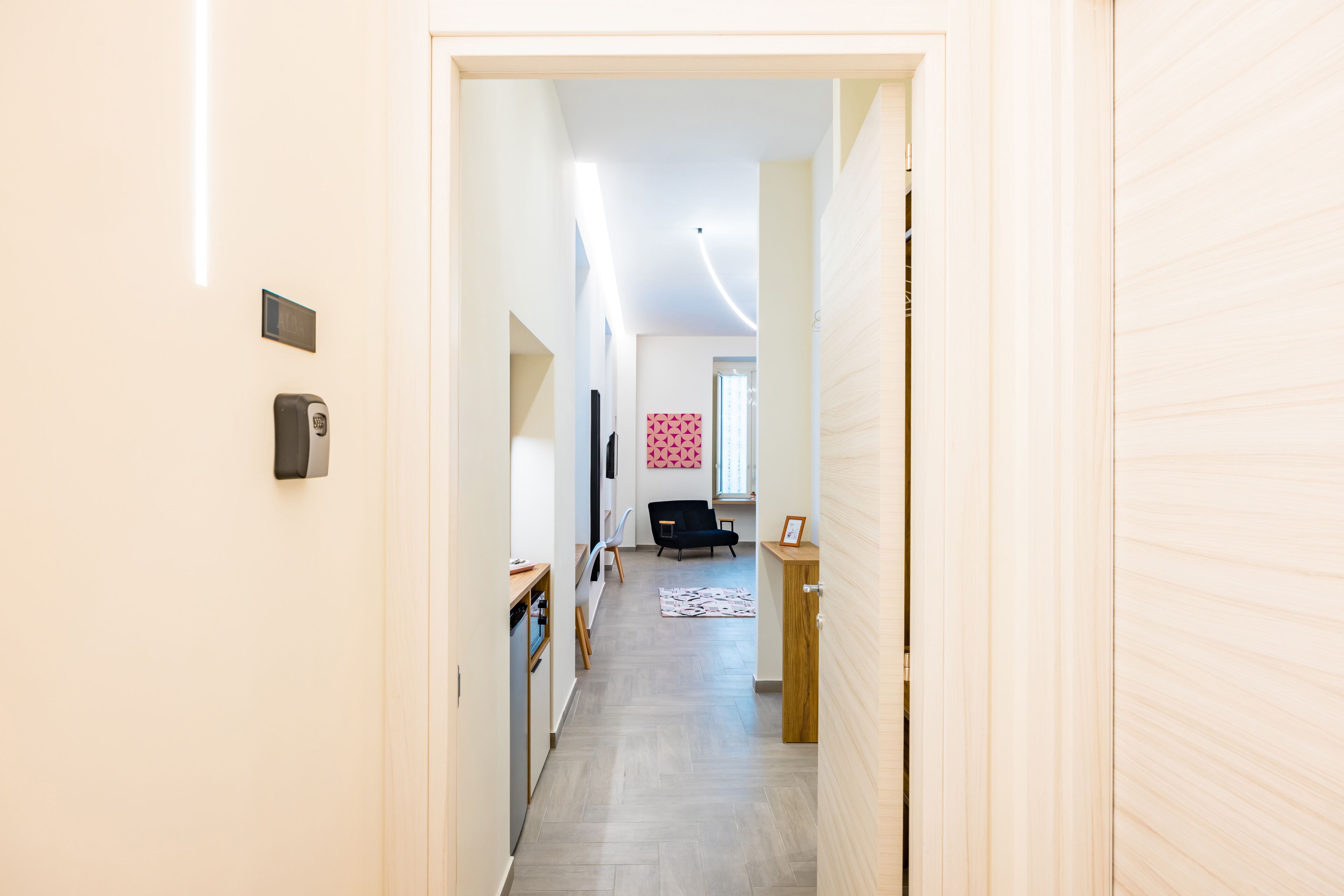 Un couloir mène à une pièce où se trouvent un lit, un bureau et une chaise. Il y a une porte sur la droite et un appareil fixé au mur sur la gauche.