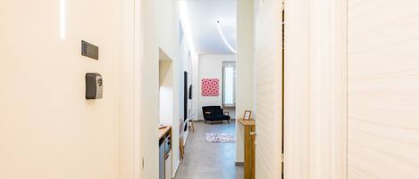 Un couloir mène à une pièce où se trouvent un lit, un bureau et une chaise. Il y a une porte sur la droite et un appareil fixé au mur sur la gauche.