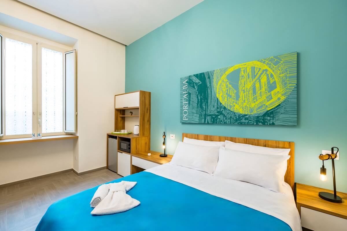 A hotel room with a bed, a desk, a chair, a window, and a wall art piece.