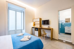 A hotel room with a bed, a desk with a TV, a chair, and a bathroom visible through an open door. - Glow Tripla - Solaris Suites (Naples)
