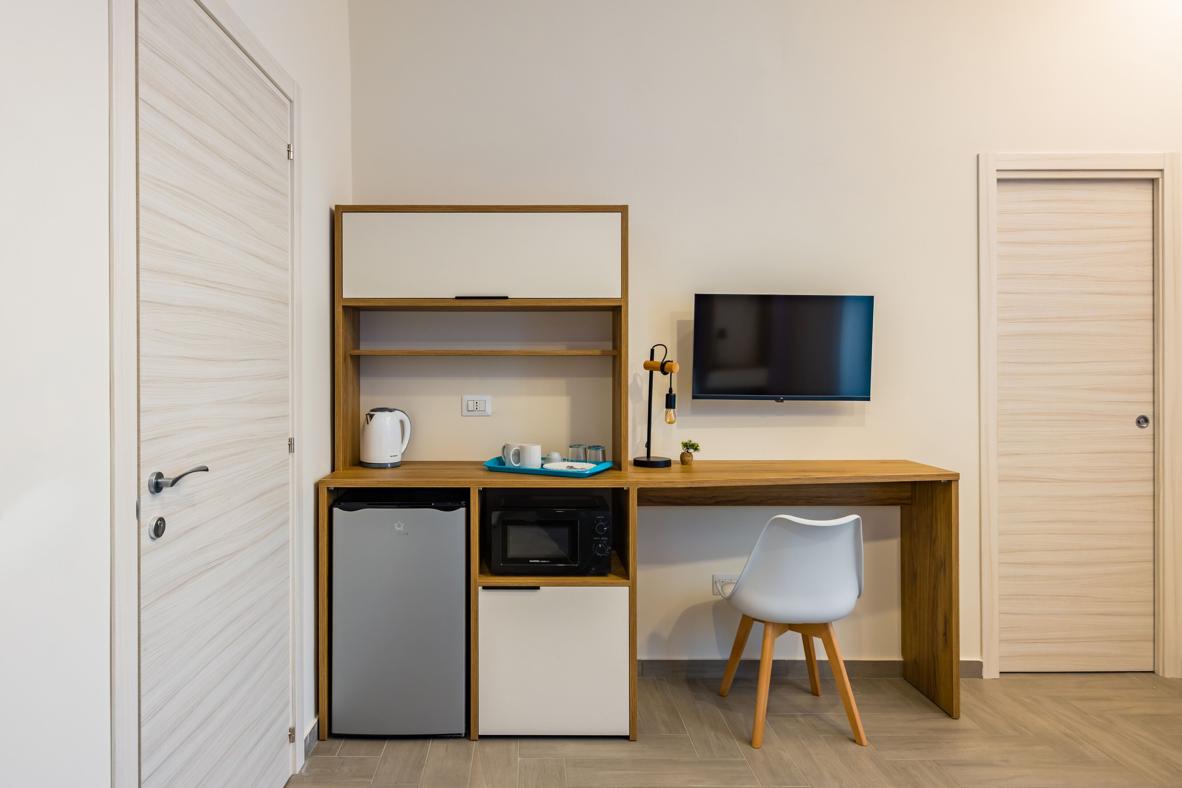 A compact kitchenette with a microwave, refrigerator, and coffee maker, a wooden desk with a chair, and a TV mounted on the wall.