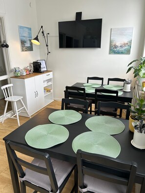Un comedor con una mesa negra, cuatro sillas, individuales verdes, un televisor montado en la pared y una planta en maceta.