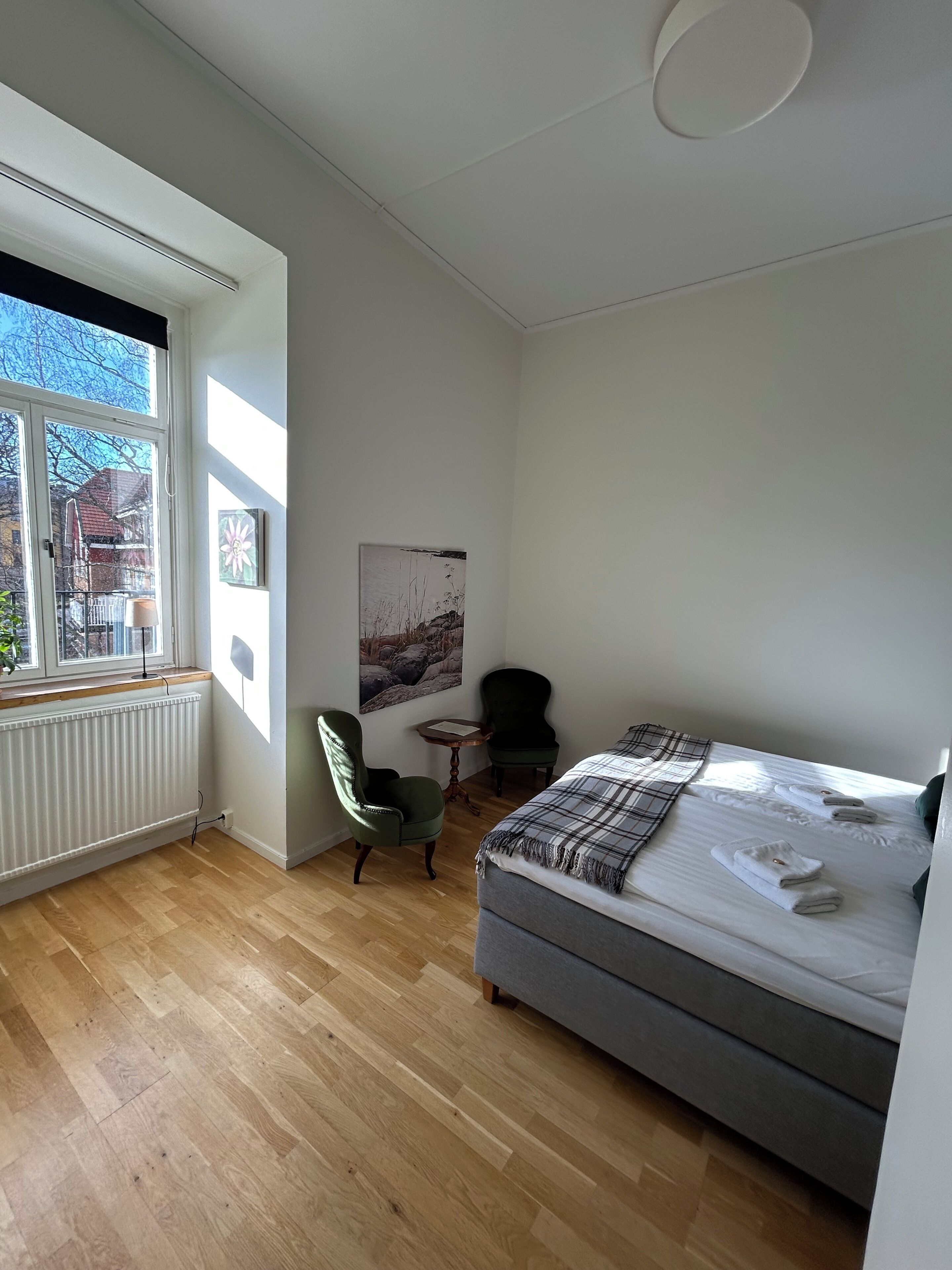 A bedroom with a bed, a chair, a small table, a radiator, and a window with a view of buildings.