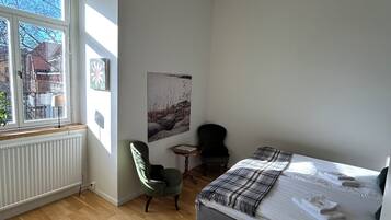 A bedroom with a bed, a chair, a small table, a radiator, and a window with a view of buildings.