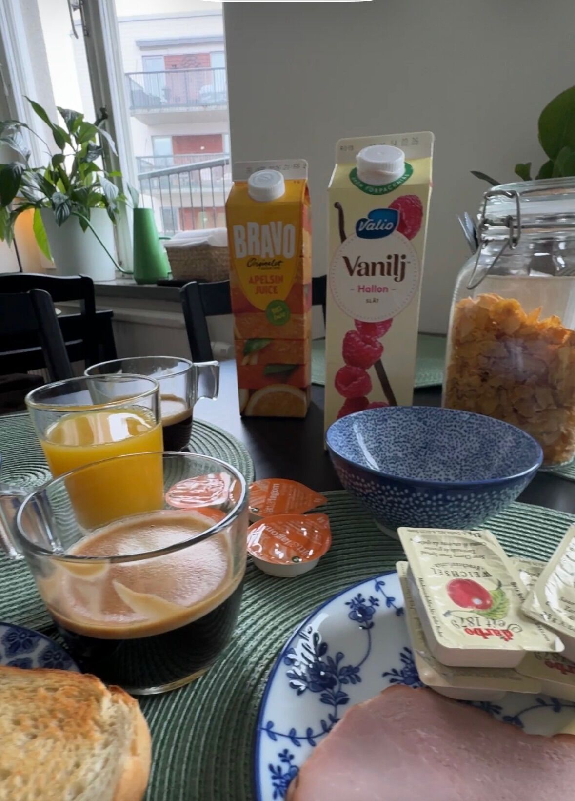 A breakfast table with a glass of orange juice, a carton of BRAVO juice, a carton of Valio vanilla milk, a glass of coffee, a bowl of cereal, a jar of jam, and a plate with a slice of ham.
