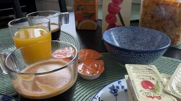 A breakfast table with a glass of orange juice, a carton of BRAVO juice, a carton of Valio vanilla milk, a glass of coffee, a bowl of cereal, a jar of jam, and a plate with a slice of ham.