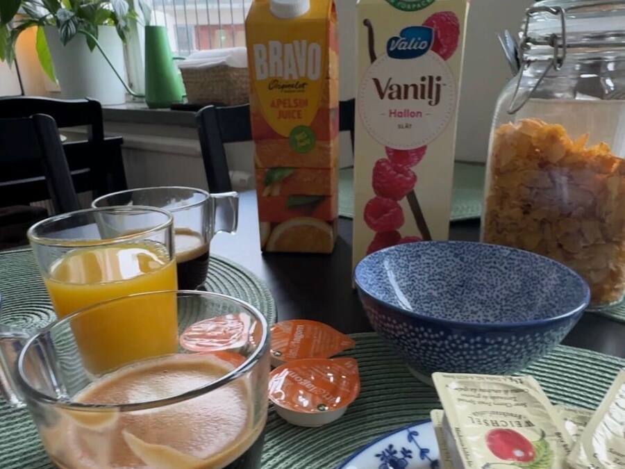 A breakfast table with a glass of orange juice, a carton of BRAVO juice, a carton of Valio vanilla milk, a glass of coffee, a bowl of cereal, a jar of jam, and a plate with a slice of ham.
