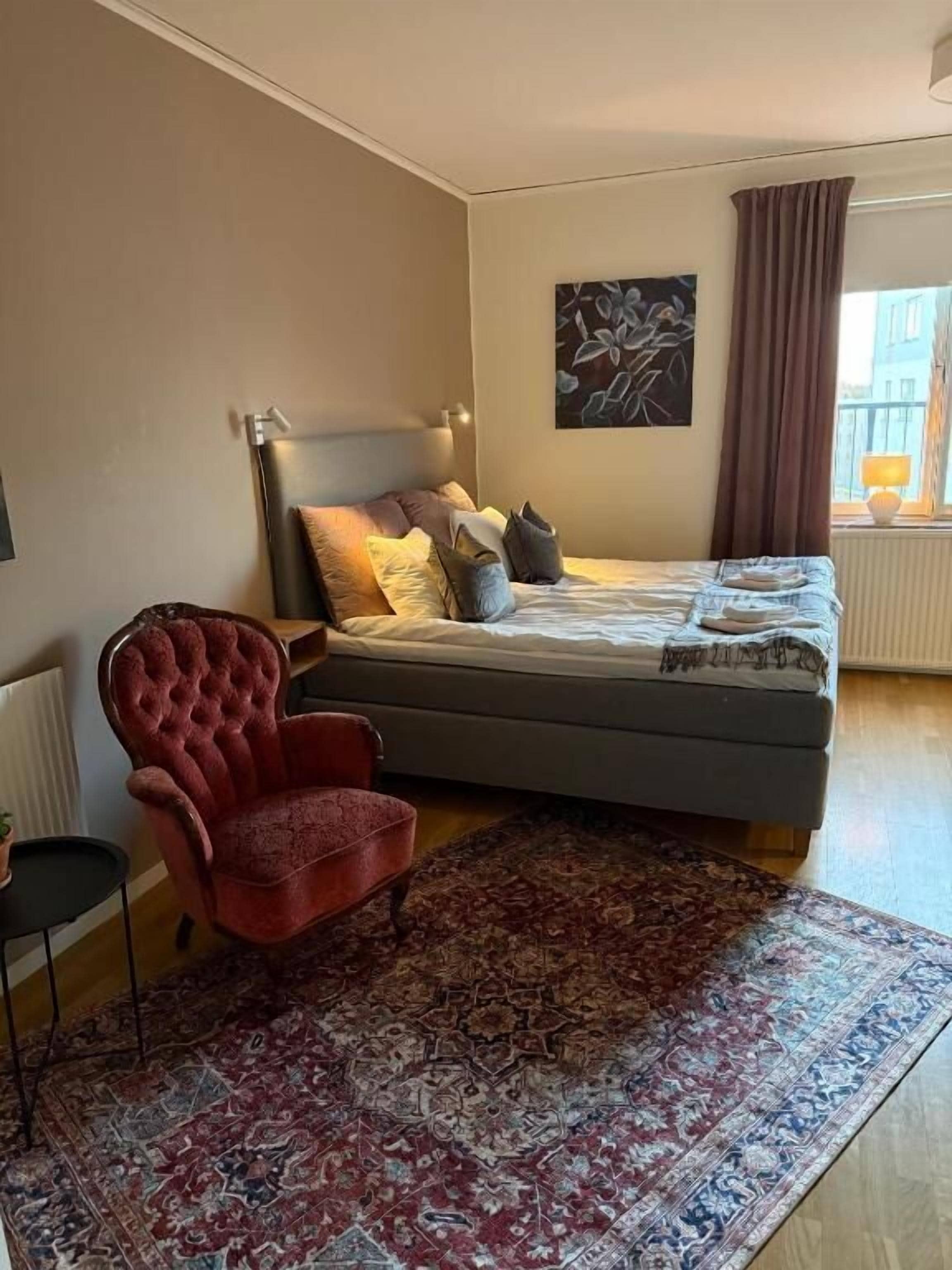 Un dormitorio con una cama, un sillón rojo, una alfombra estampada, un cuadro en la pared y una ventana con cortinas.