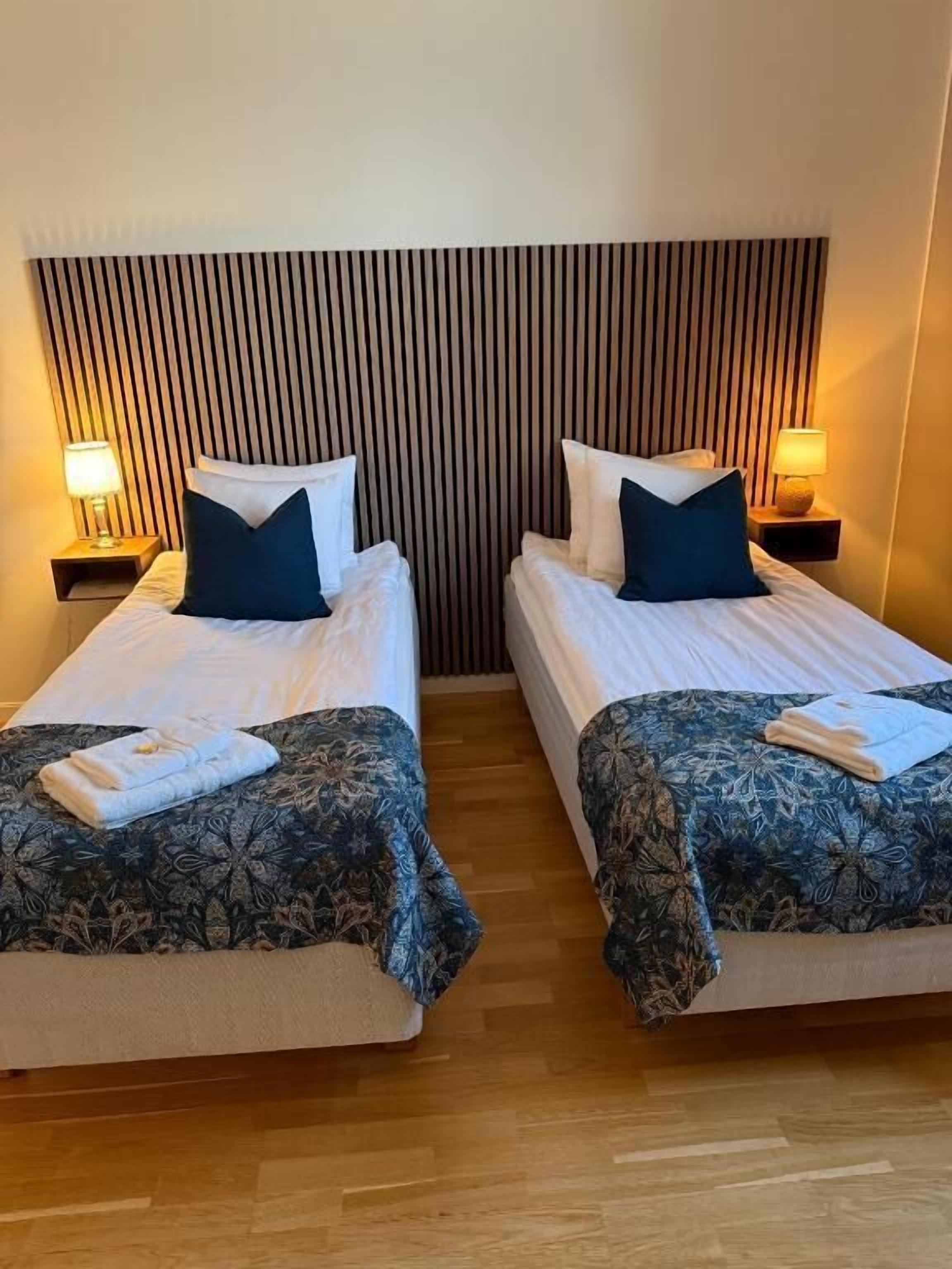 A hotel room with two beds, each with a patterned bedspread, pillows, and a nightstand with a lamp.