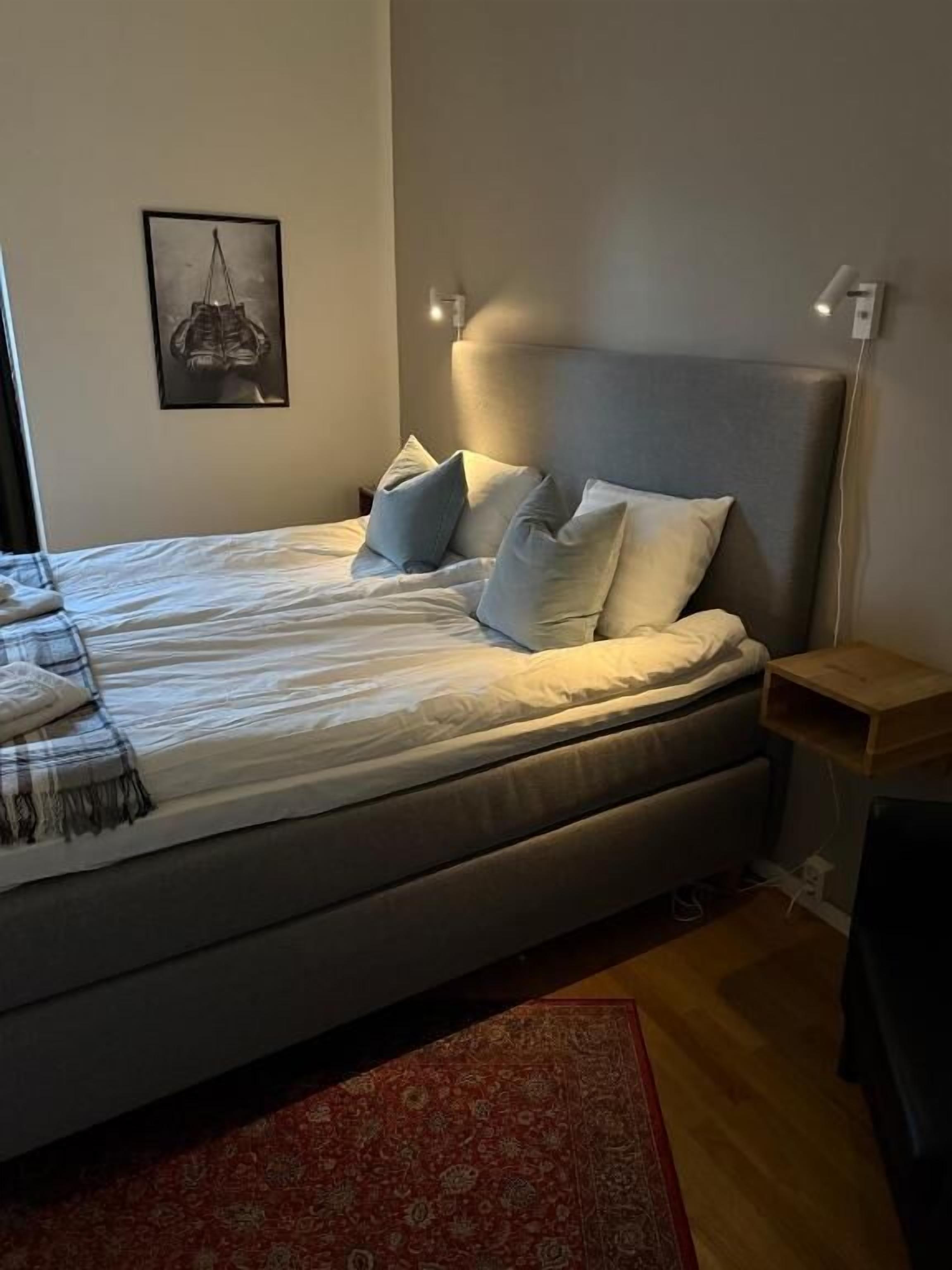 Un dormitorio con cama, almohadas, mesita de noche, luz empotrada en la pared y un cuadro enmarcado.