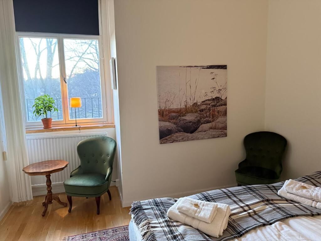 Un dormitorio con una cama, dos sillas, una mesita, una ventana con vistas a árboles y un cuadro en la pared.