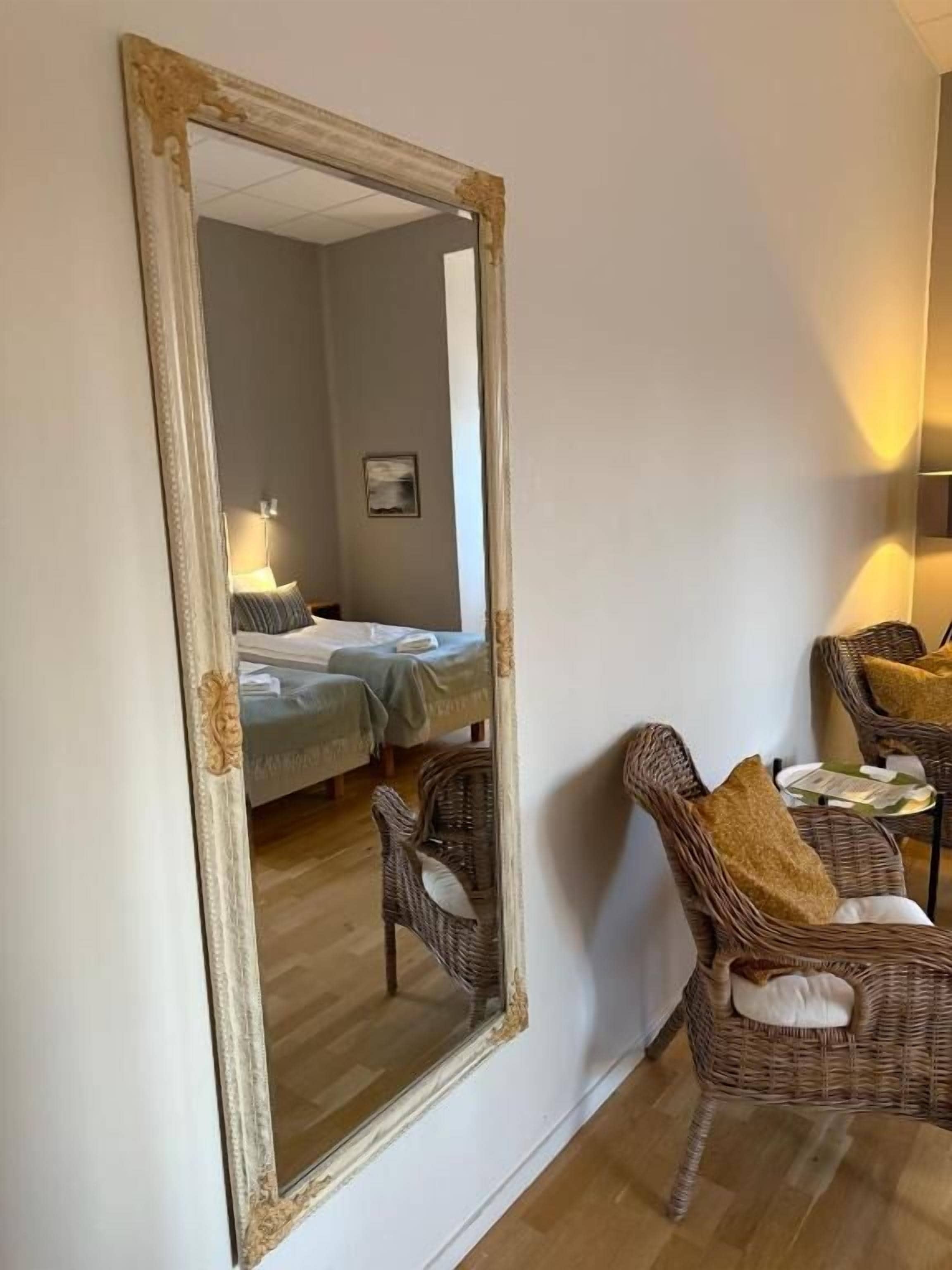 A room with a large mirror, wicker furniture, a bed, and a small table with a lamp.