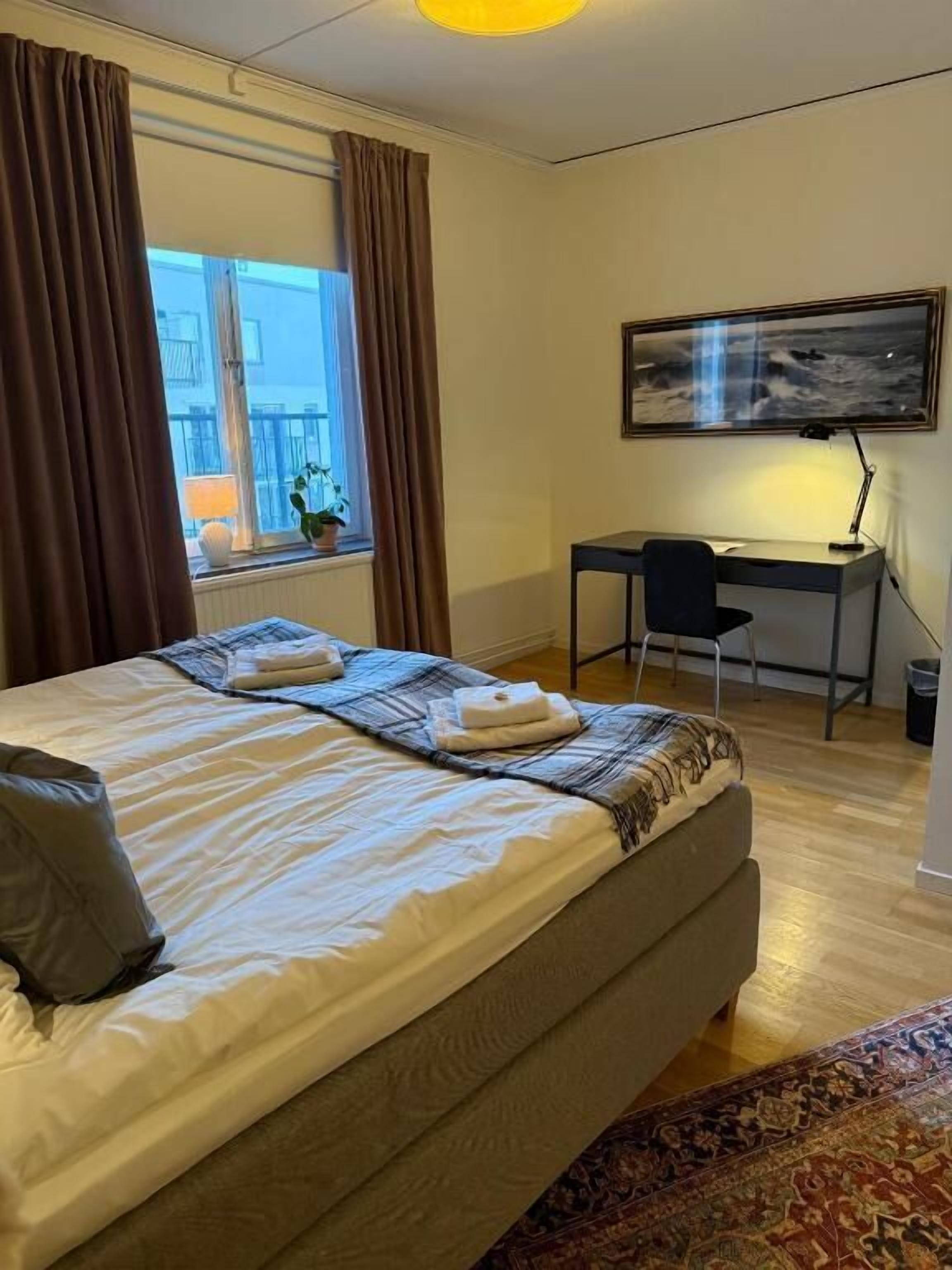 Un dormitorio con cama, escritorio, silla, ventana con cortinas y un cuadro en la pared.