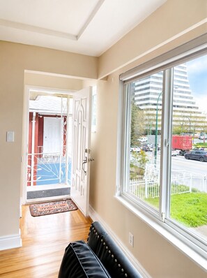 A hallway with a black leather sofa, a large window with a view of a street and buildings, and a door leading to another room. - Park View 3BR Home.Covered SunDeck. Near Metrotown (Vancouver)
