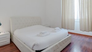 Una cama bien hecha con sábanas y toallas blancas, una mesita de noche con lámpara y una ventana con cortinas.
