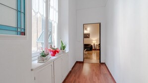 Un pasillo con piso de madera, paredes blancas, un ventanal con vidrios azules y una puerta que da a una habitación con cama y lámpara.