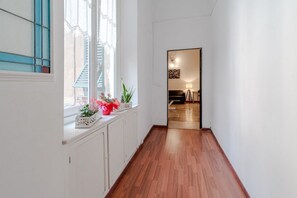 Ein Korridor mit Holzdielen, weißen Wänden, einem Fenster mit blauen Glasfeldern und einer Tür zu einem Zimmer mit einem Bett und einer Lampe.