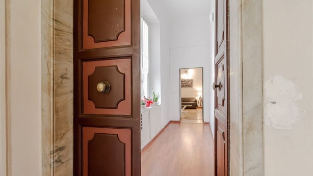 Un pasillo con puertas de madera, suelo de mármol y vista a una habitación con cama y mesita de noche.