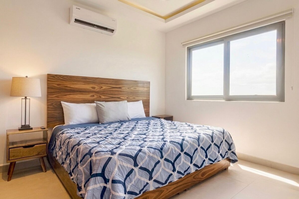 A bedroom with a wooden headboard, a bed with a patterned bedspread, a nightstand with a lamp, and a window with blinds.
