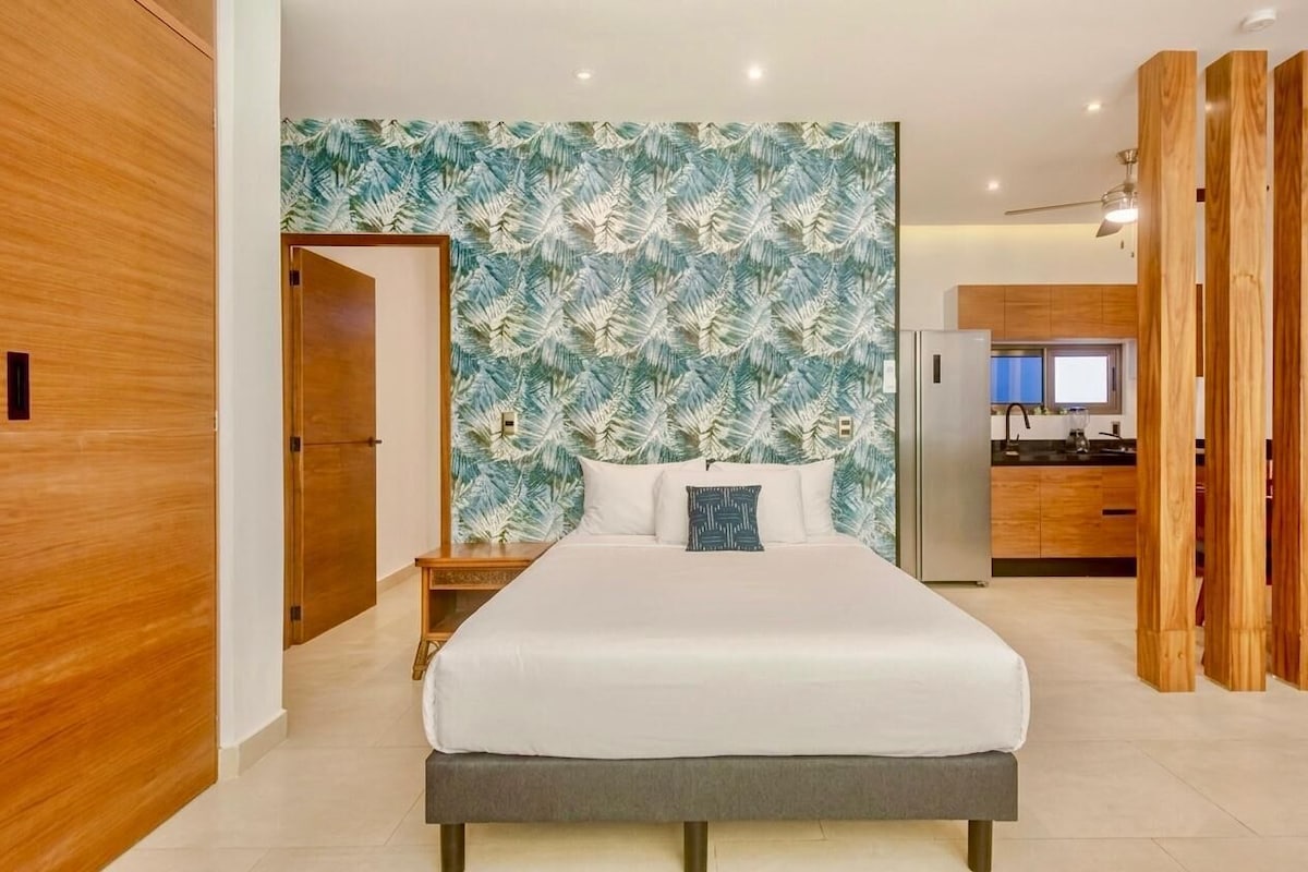 A modern bedroom with a large bed, a wooden wardrobe, and a kitchen area in the background.
