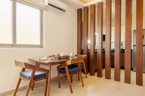 Un comedor con mesa y sillas de madera, una cocina al fondo y una ventana con equipo de aire acondicionado.