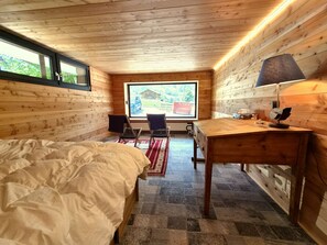 Ein Zimmer in einer Holzhütte mit Bett, Schreibtisch, Stühlen und einem großen Fenster.