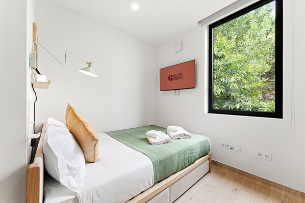 Een moderne hotelkamer met een bed, een televisie, een raam met uitzicht op groen en een wandlamp.