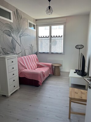 Ein Zimmer mit einem gestreiften Sofa, einem weißen Schminktisch, einem kleinen runden Tisch, einem Fernseher auf einem Holzständer und einem großen Fenster mit transparenten Vorhängen.