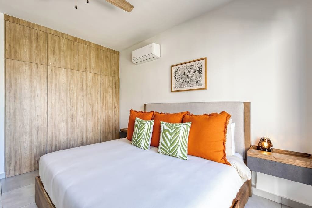 A bedroom with a wooden panel wall, a bed with white and orange bedding, a framed picture on the wall, and a nightstand with a lamp.