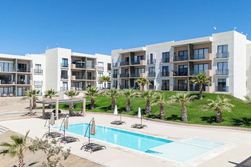 A modern hotel with a swimming pool, sun loungers, and palm trees.