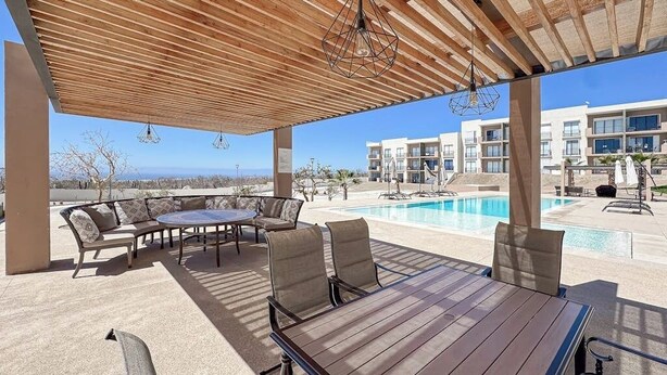 An outdoor patio with a wooden table and chairs, a pool, and a building in the background. - Bright and Modern Condo w/ Balcony, Shared Pool, A/c, Minutes to Beach & Town (Cabo San Lucas)