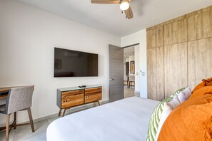 A modern bedroom with a large bed, a desk with a chair, a TV mounted on the wall, and a wooden panel wall. - Bright and Modern Condo w/ Balcony, Shared Pool, A/c, Minutes to Beach & Town (Cabo San Lucas)