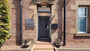 A stone building with a dark entrance door, a welcome sign, and two flower pots on the ground. - Winmar Guesthouse (Inverness)
