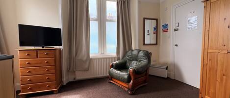 A room with a large window, a leather armchair, a wooden dresser, a television, and a wardrobe.