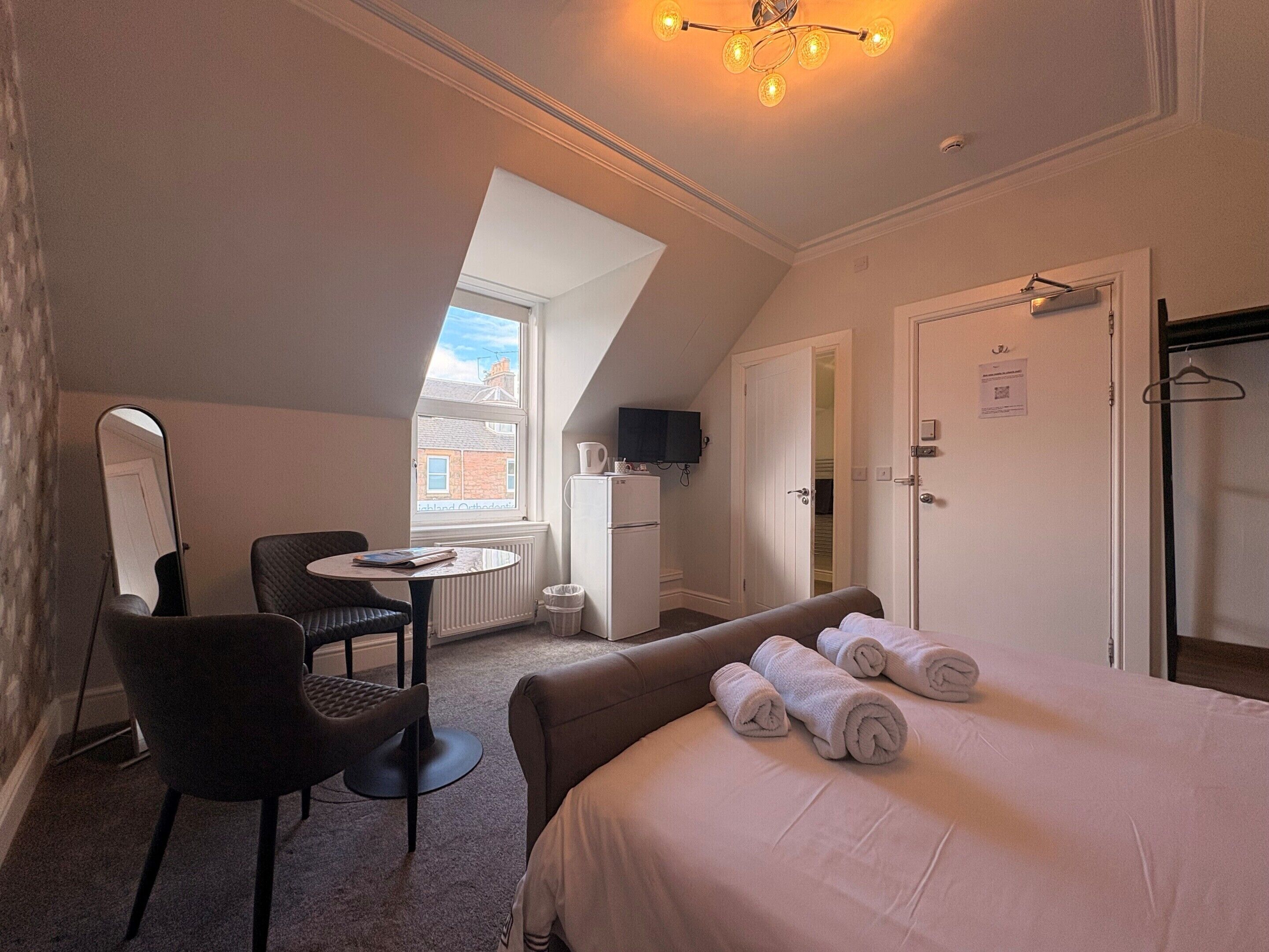 A compact hotel room with a bed, a chair, a small table, a mirror, a television, and a window.